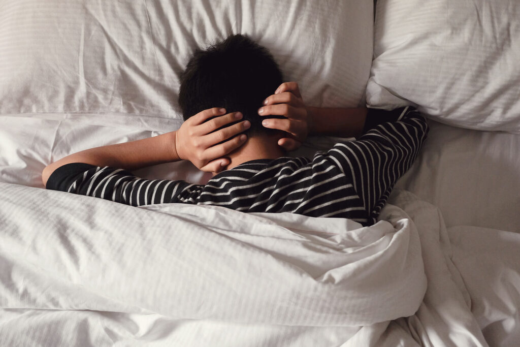 Young man in bed with his hands over his ears representing someone that is overwhelmed by social anxiety and ADHD. Regain control of your life with ADHD-Focused Therapy for Anxiety in NY, NY.