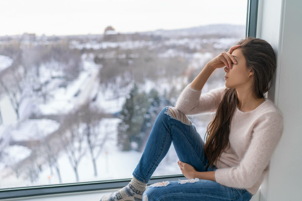 Young woman struggling with anxious thoughts sits alone looking out the window. You are not in this alone, find the help you need with ADHD-Focused Therapy for Anxiety in NYC.