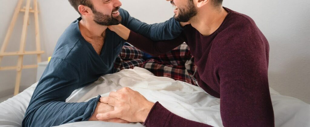 Gay couple sitting together on a bed laughing and talking representing the improved communication and bond that can be created with the help of ADHD-Focused Couples Therapy in NYC.