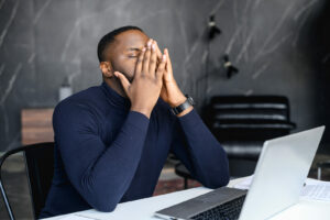 A man covers his face while sitting at his computer desk. Learn more about ADHD therapy in NYC and the support it can offer to help manage ADHD without medication today. 
