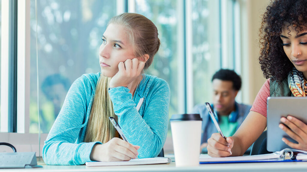 A picture of a teen staring out the window. She is seated next to a student looking at her computer and writing.