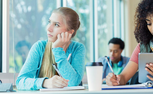 A picture of a teen staring out the window. She is seated next to a student looking at her computer and writing.